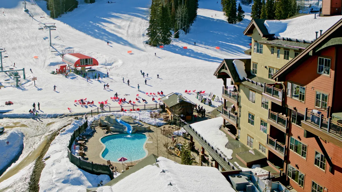 A dynamic exterior view featuring the lodge with ski slopes just steps away, a heated swimming pool, and the ski lift in the background. The image is lively, showing guests relaxing by the pool after a day on the slopes while others continue to ski and snowboard, capturing both excitement and relaxation in one frame.