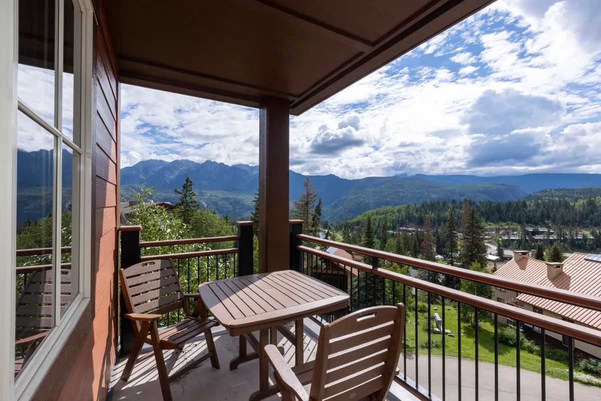 The image shows a condo balcony with comfortable outdoor furniture overlooking majestic mountain peaks, bathed in the warm glow of sunset. The sky is vivid, with hues of orange and purple, and the scene evokes a peaceful, relaxing atmosphere perfect for unwinding or enjoying a meal with a view.