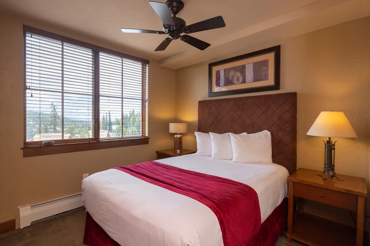 A restful bedroom with a bed adorned with a red throw, large windows that offer scenic outdoor views, a decorative lamp, and framed artwork. The tranquil atmosphere is perfect for unwinding and enjoying fresh mountain breezes.
