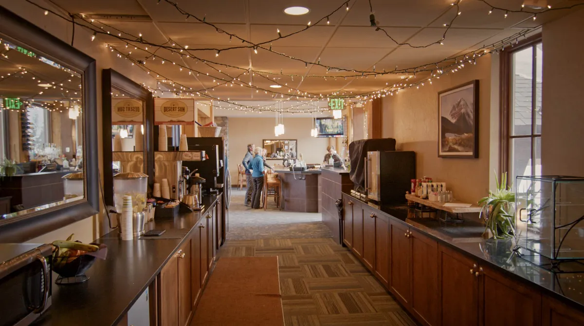 A warm and inviting interior bar area, with string lights overhead, a wooden counter, and a cozy coffee station. The atmosphere feels intimate—ideal for gathering, socializing, and enjoying food and drinks together.