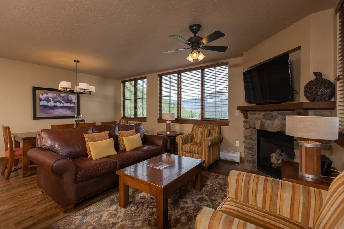 A warm and expansive open-plan living and dining area featuring a plush leather sectional, a rustic wooden coffee table, a prominent stone gas fireplace, and large windows that frame breathtaking mountain views. The dining table is positioned to encourage communal meals and gatherings, while the flat-screen TV provides entertainment for cozy nights in.