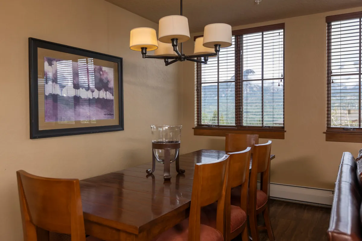 A dining area featuring a large table under a chandelier, artwork on the wall, and expansive windows that flood the space with natural light. The windows frame views of the outdoors and create an airy, bright atmosphere.