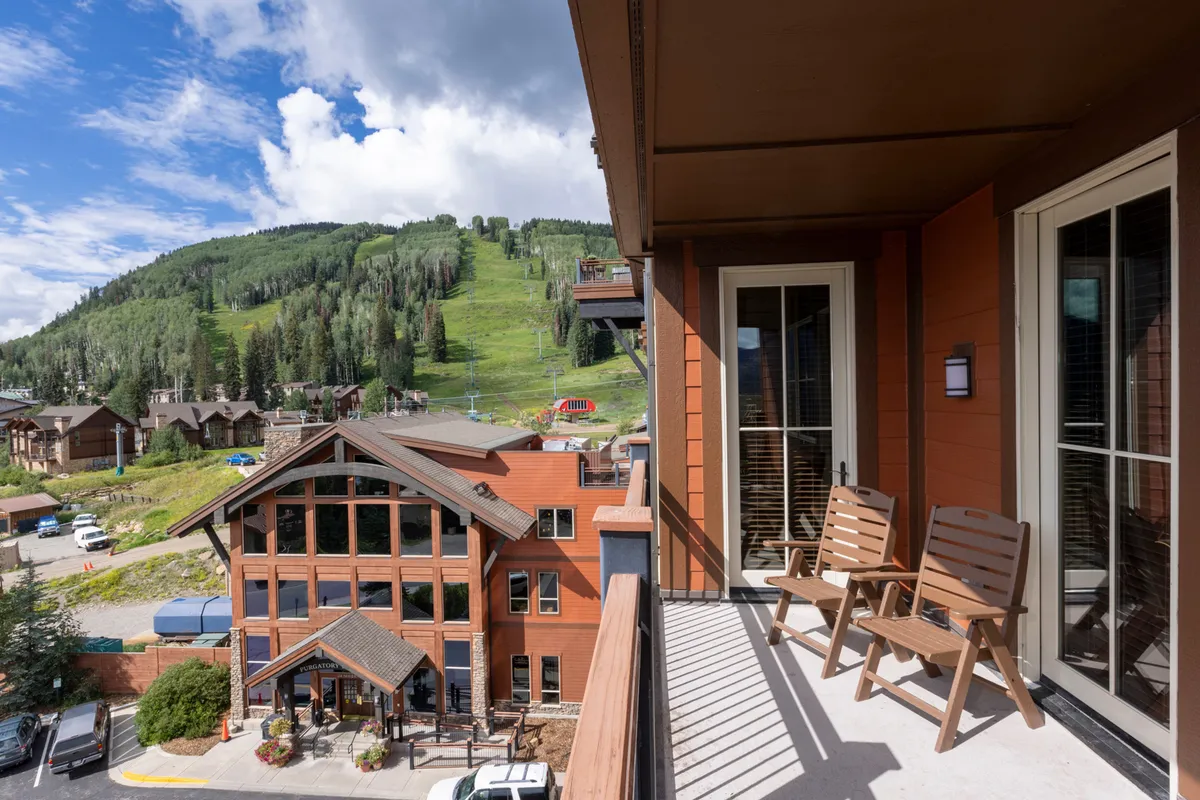 A tranquil scene showing a resort balcony with comfortable seating overlooking the beautiful mountain landscape and the surrounding resort buildings. The soft hues of sunset set a relaxing tone, inviting families to unwind while enjoying the open air and scenic views.