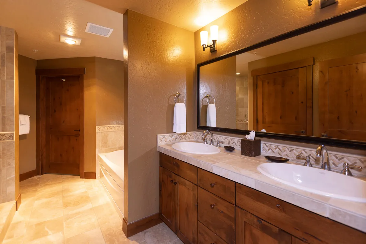 A spacious, serene bathroom with a double sink vanity, large mirror, wooden cabinetry, and a bathtub. The space is well-lit and designed for relaxation and self-care.