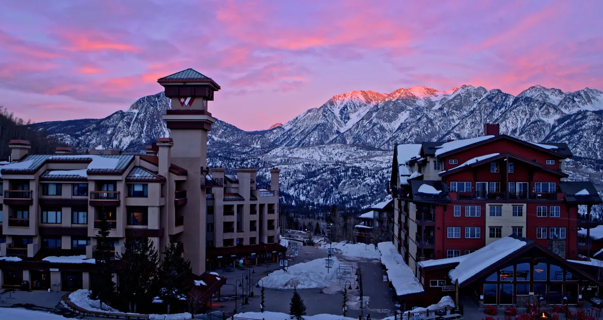 This exterior image captures stunning snow-covered mountains as the backdrop, with resort buildings nestled in the landscape and a serene sky overhead. The tranquil scene suggests both adventure and relaxation, with winter sports activities like skiing and snowboarding implied, while also evoking the romantic ambiance of a mountain retreat.