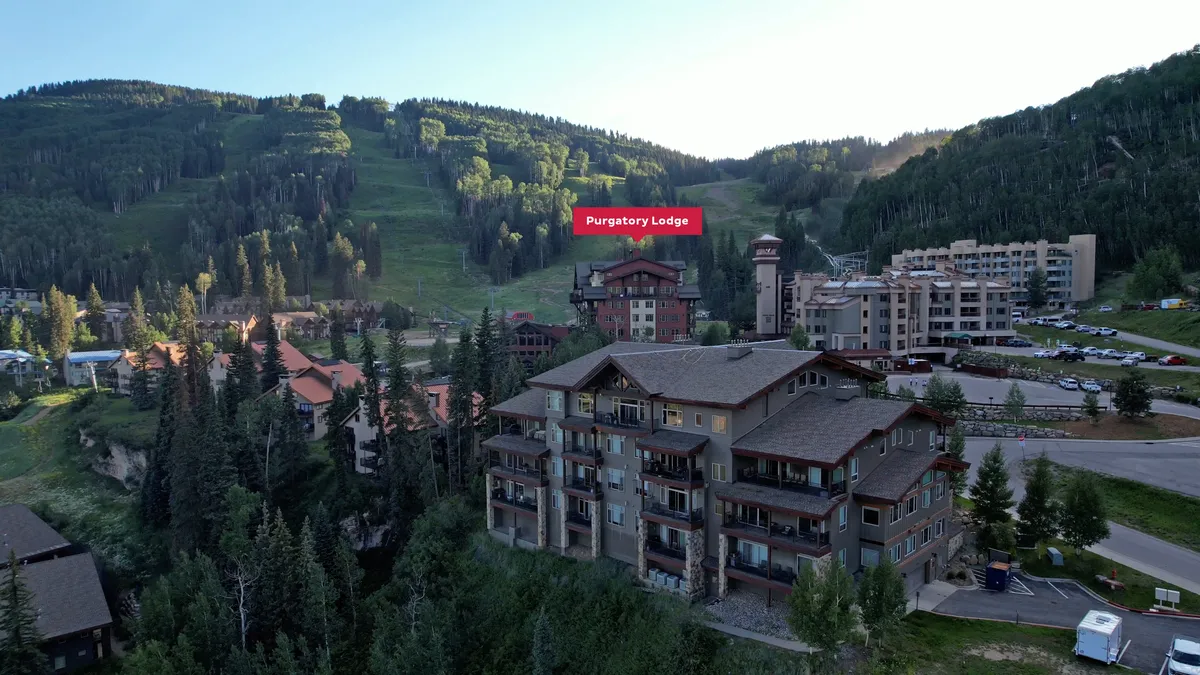 A tranquil exterior shot of Purgatory Lodge with the mountains rising behind, nearby buildings, and hints of village life. The image suggests easy access to both the slopes and the vibrant heart of Purgatory Village, with visible walkways and a welcoming ambiance for families and groups.