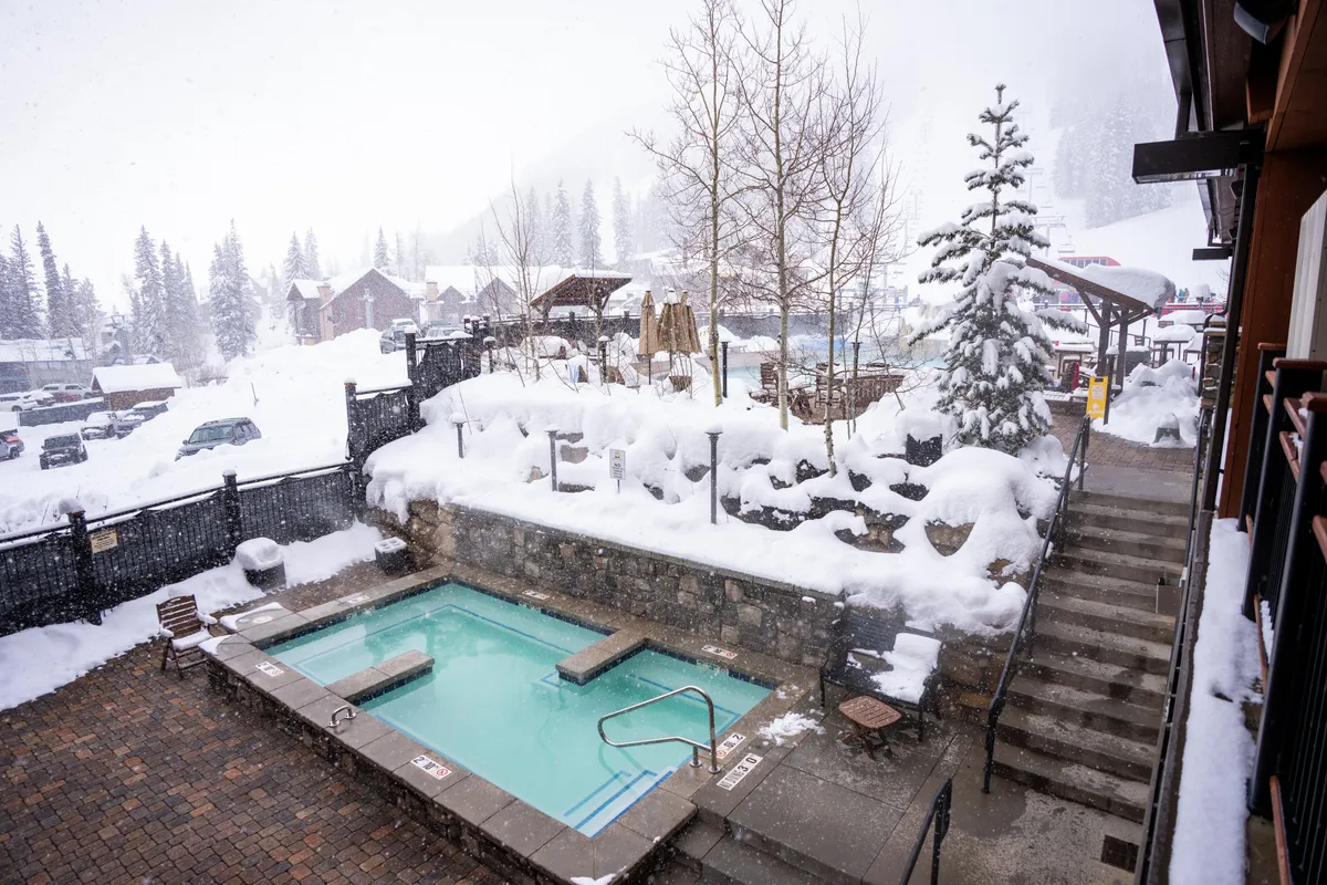 This image presents an outdoor hot tub (swim spa) set against a snow-covered landscape, with trees and steps leading up. The scene is serene, evoking relaxation and warmth, even in a winter setting.
