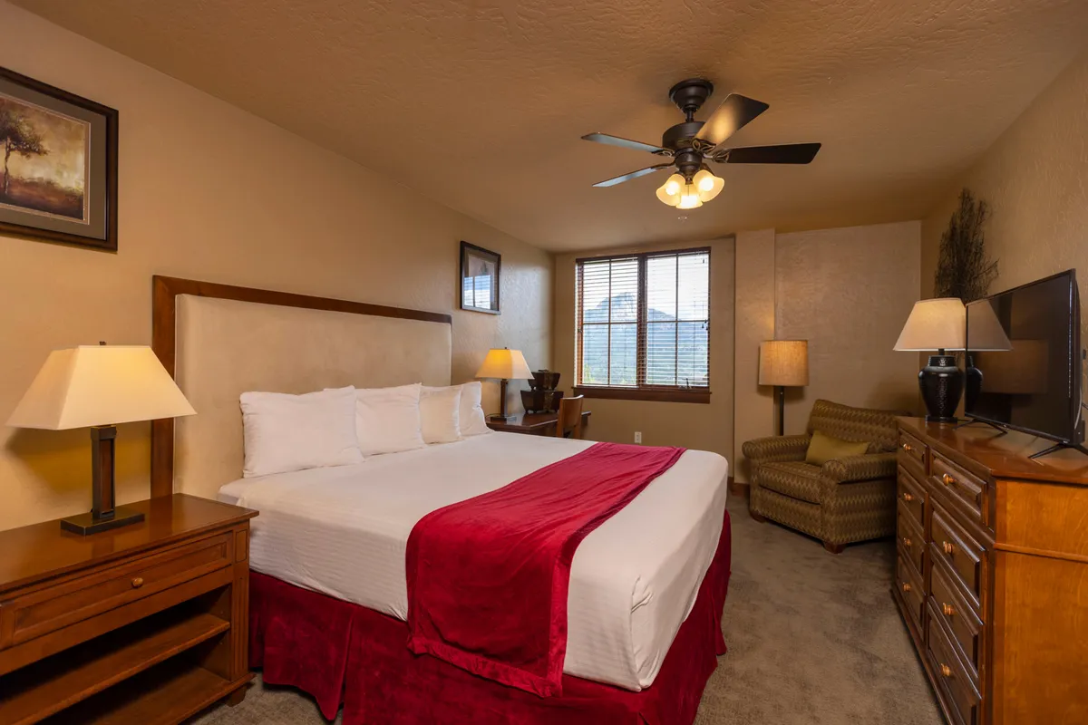 A warmly lit bedroom with a bed featuring a red throw, wooden furniture, a TV, and a seating area. Large windows fill the room with natural light, and the decor exudes comfort and relaxation.