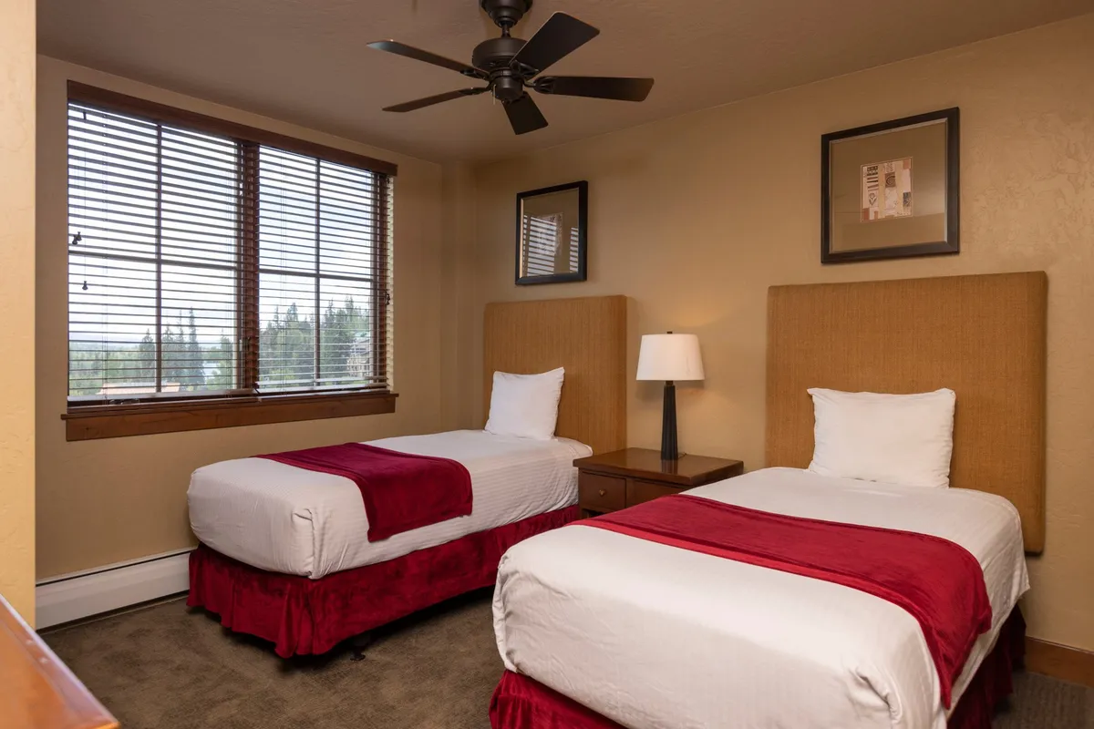 A bright bedroom with two twin beds, a table lamp between them, a large window with blinds, and decorative wall art. The layout is ideal for families or groups, offering flexibility and privacy.