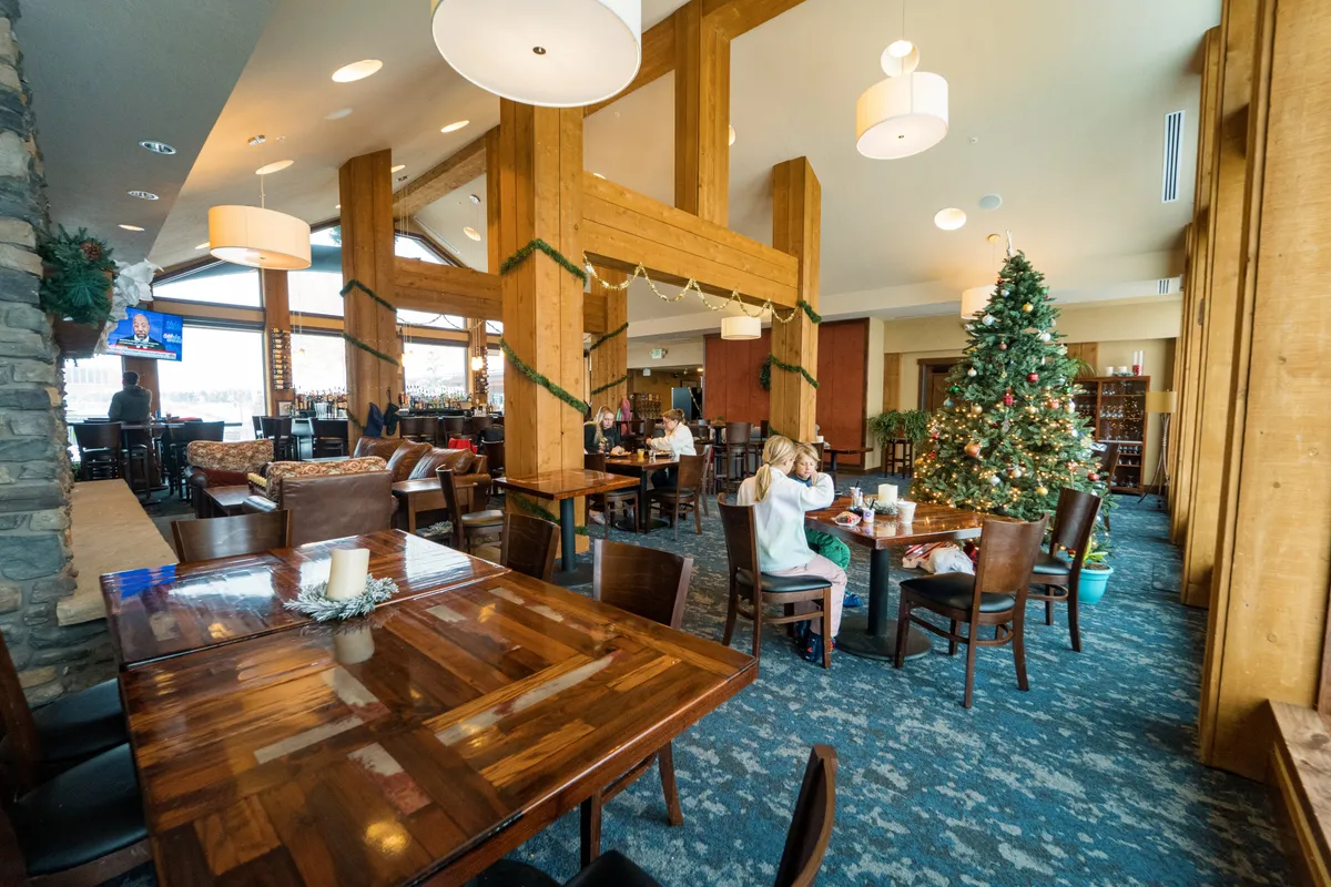 A festive interior scene with a decorated Christmas tree, wooden beams, communal dining tables, and a bar area, radiating warmth and joyful holiday spirit. The space feels welcoming for group gatherings, celebrations, and cozy evenings together.